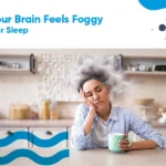 Woman experiencing brain fog and fatigue after poor sleep, sitting at a kitchen counter with a mug.